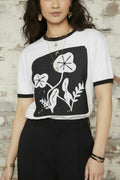 A woman modeling a white ringer t-shirt with black trim and a black square graphic featuring white stylized flowers, paired with black jeans against a plain white background.