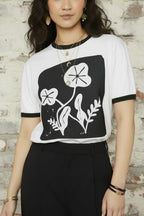 A woman modeling a white ringer t-shirt with black trim and a black square graphic featuring white stylized flowers, paired with black jeans against a plain white background.