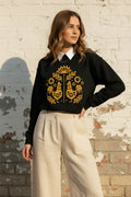 A woman wearing a black crewneck sweatshirt with a yellow folk-art graphic of two roosters and flowers, paired with gray denim shorts and holding an iced coffee.