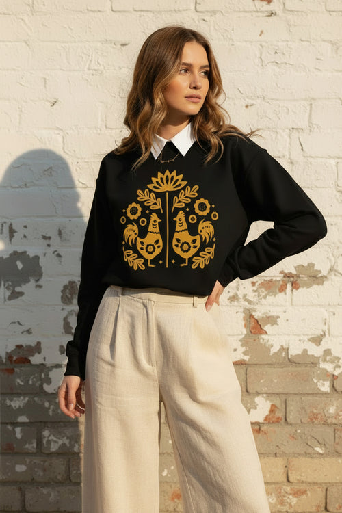 A woman wearing a black crewneck sweatshirt with a yellow folk-art graphic of two roosters and flowers, paired with gray denim shorts and holding an iced coffee.