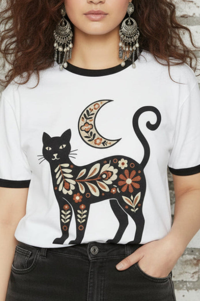 A woman modeling a white ringer t-shirt with black trim and a graphic of a black cat decorated with orange, yellow, and gold patterns next to a crescent moon, paired with black jeans against a plain white background.