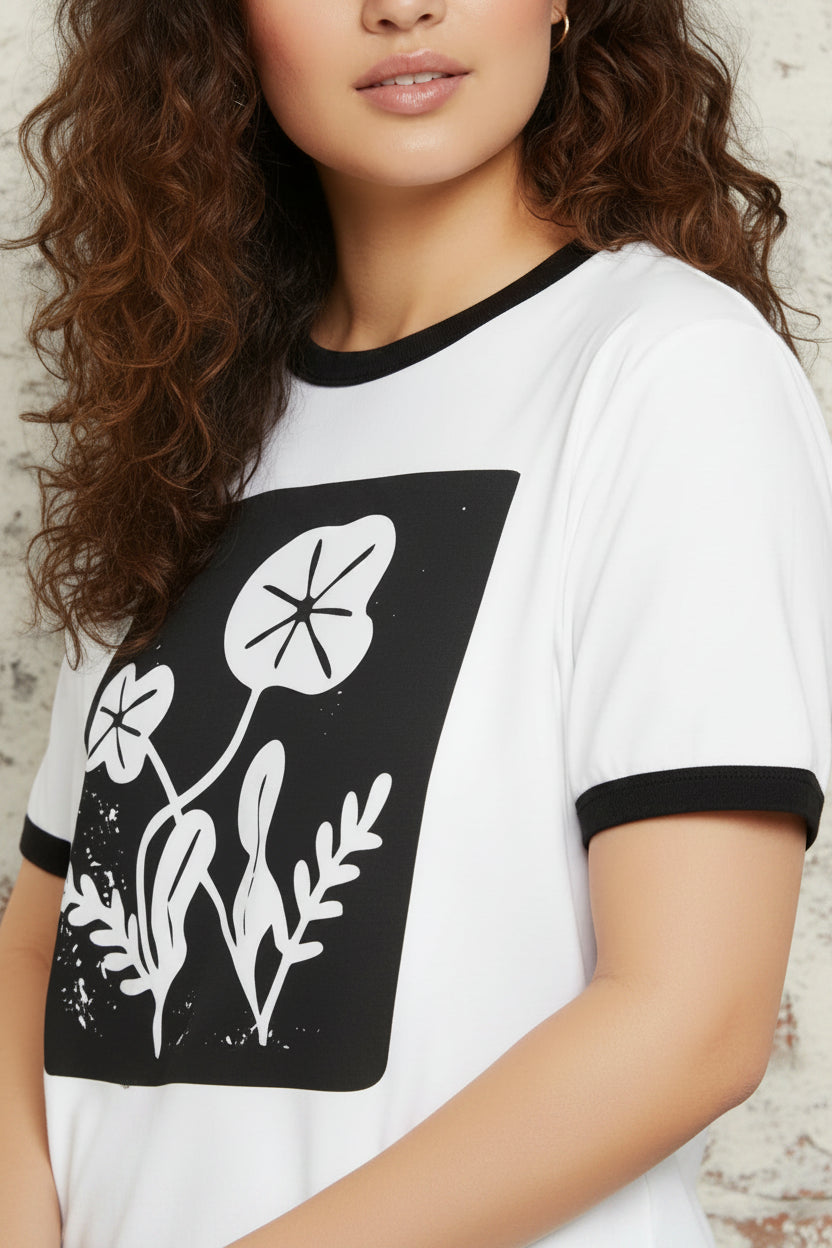 A woman wearing a white ringer T-shirt with black trim at the collar and sleeve cuffs, featuring a large black graphic panel on the front with white botanical leaf illustrations.