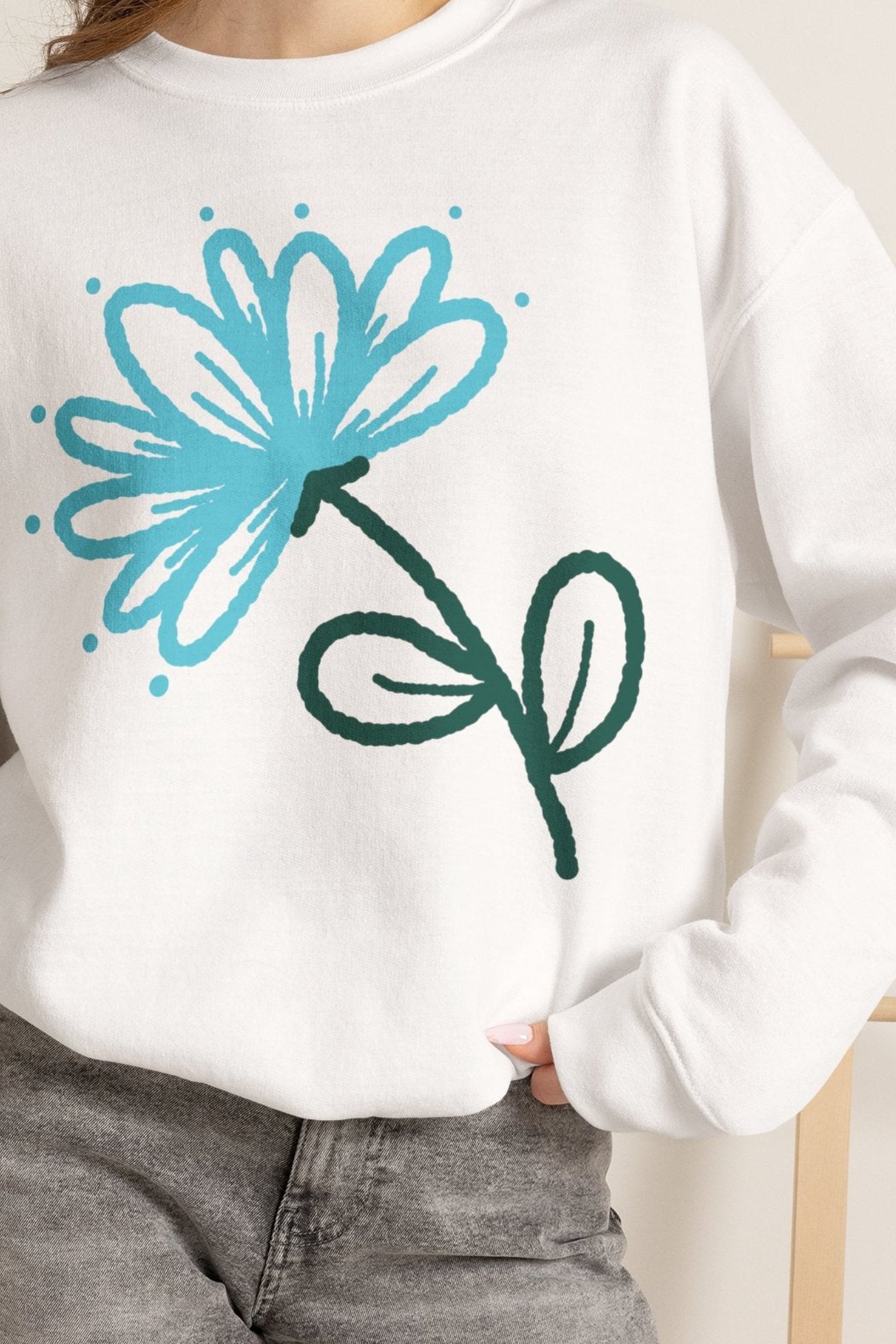 A woman wearing a white crewneck sweatshirt with a blue and green flower graphic, paired with gray denim shorts and holding an iced coffee.