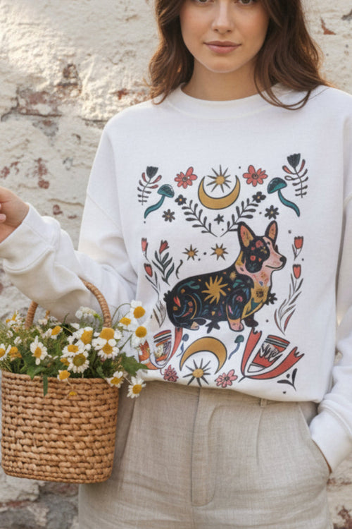 A woman wearing a white crewneck sweatshirt with a colorful, detailed graphic of a corgi surrounded by celestial and floral motifs. She is holding an iced coffee and wearing gray denim shorts in a brightly lit indoor setting.
