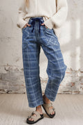 A person wearing loose blue plaid pants with a dark blue drawstring waist, a gray knit sweater, and beige sandals, standing against a plain light wall and textured floor.