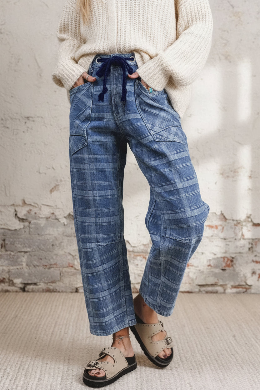 A person wearing loose blue plaid pants with a dark blue drawstring waist, a gray knit sweater, and beige sandals, standing against a plain light wall and textured floor.