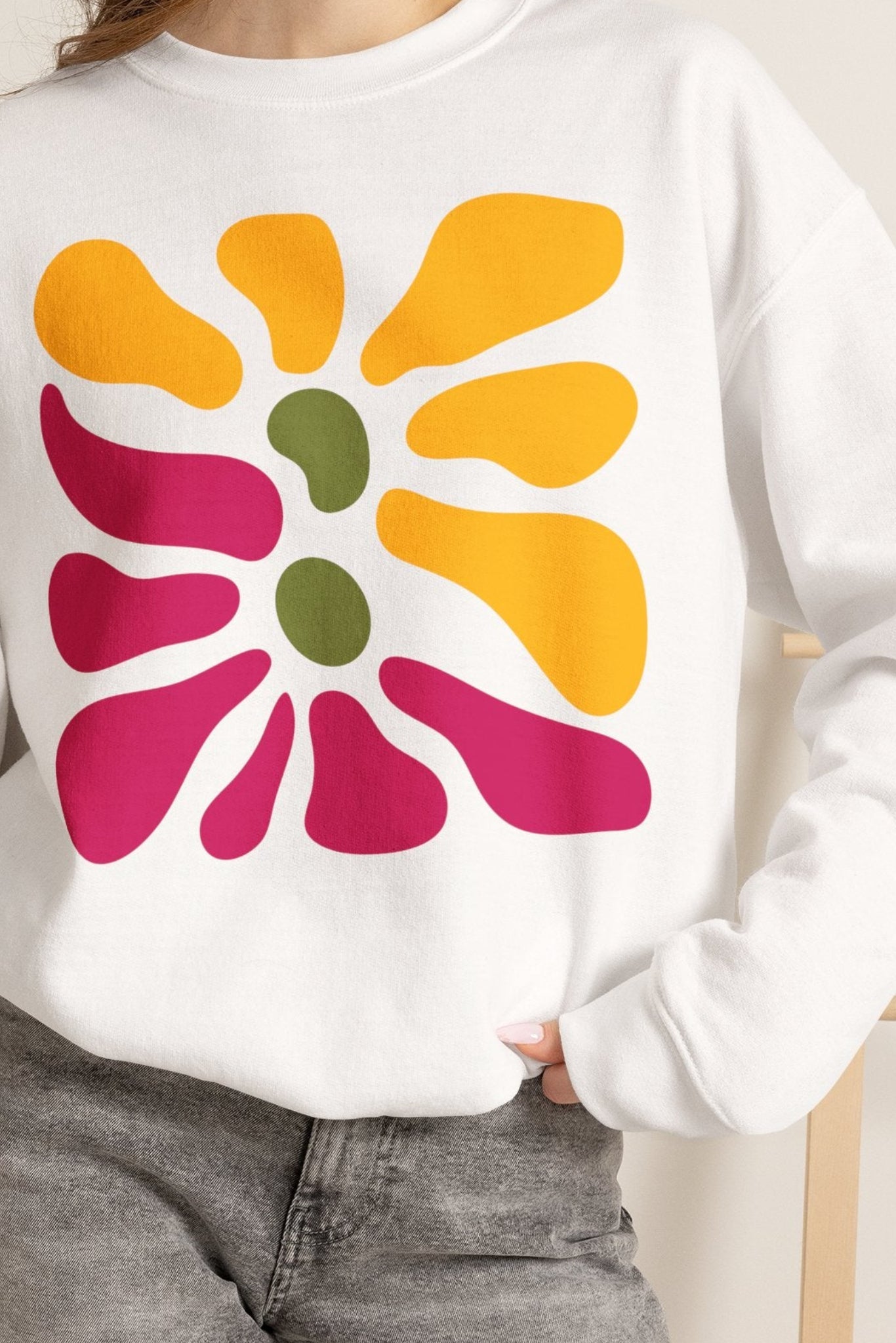 A woman wearing a white crewneck sweatshirt with a retro 1960s-style flower graphic in yellow, magenta, and green, paired with gray denim shorts and holding an iced coffee.