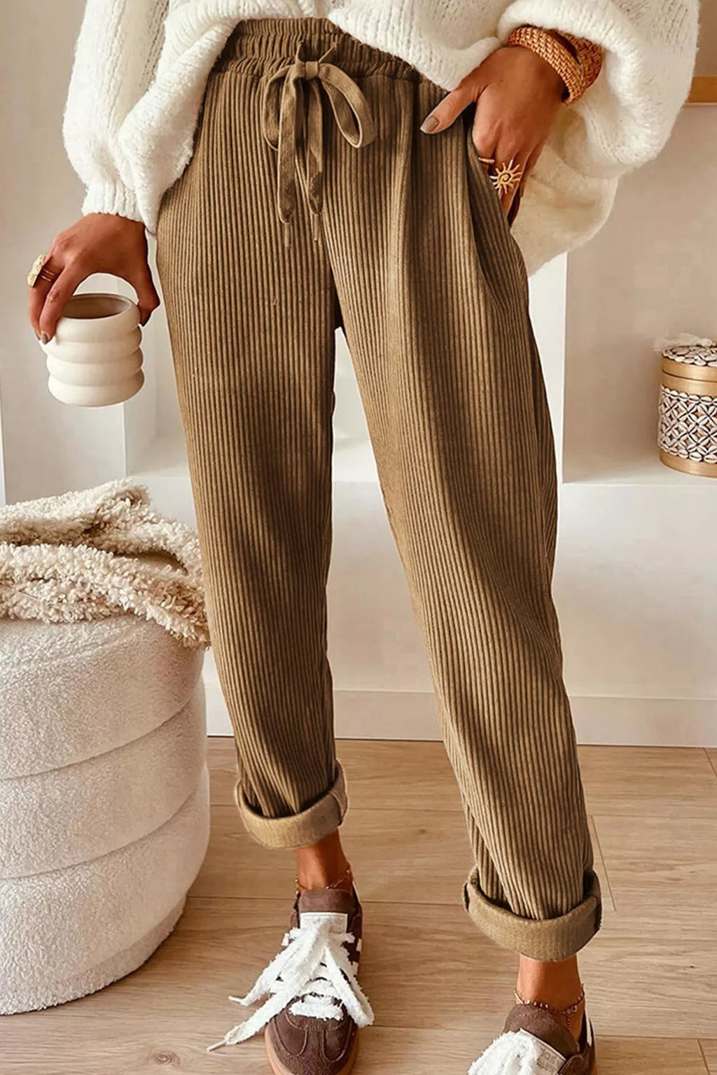 Person wearing brown ribbed pants with a white top and sneakers, holding a ceramic cup.