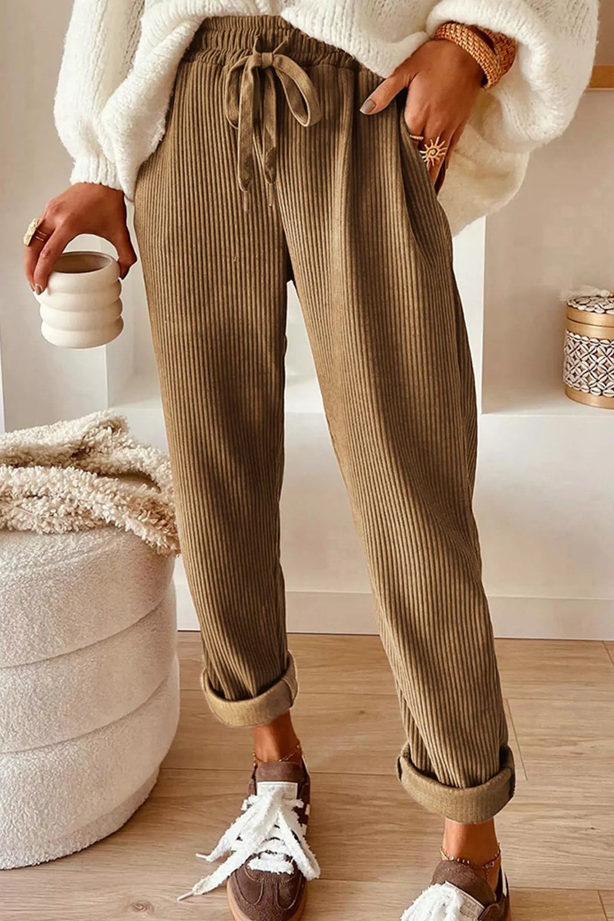 Person wearing brown ribbed pants with a white top and sneakers, holding a ceramic cup.