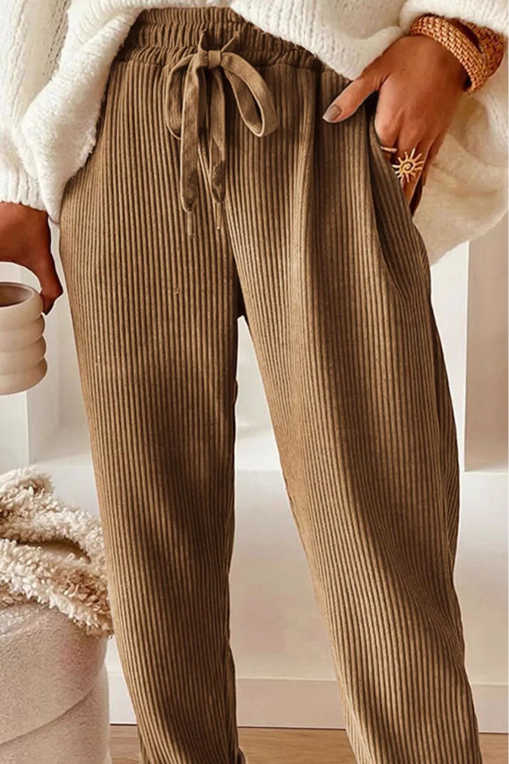 Person wearing brown ribbed pants holding a white mug indoors.