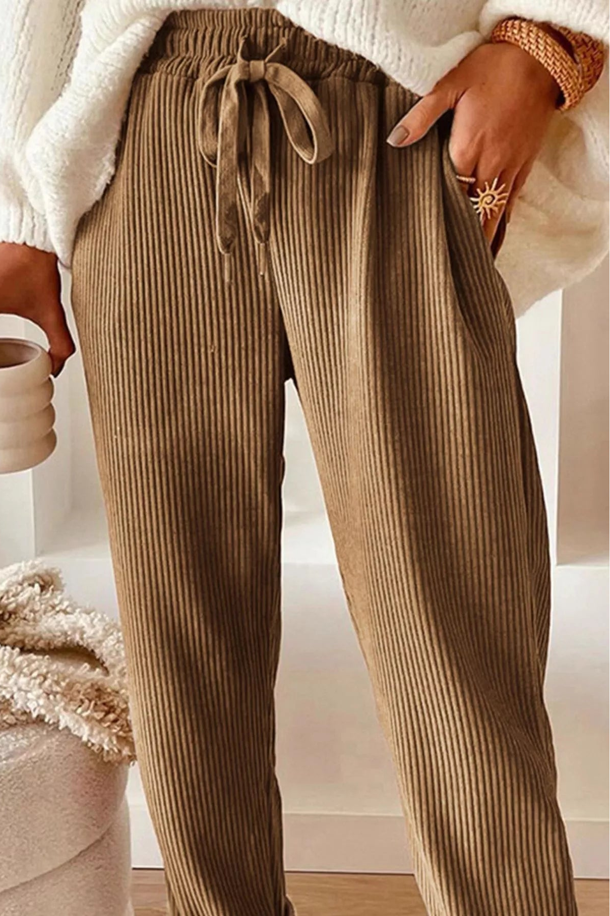 Person wearing brown ribbed pants holding a white mug indoors.