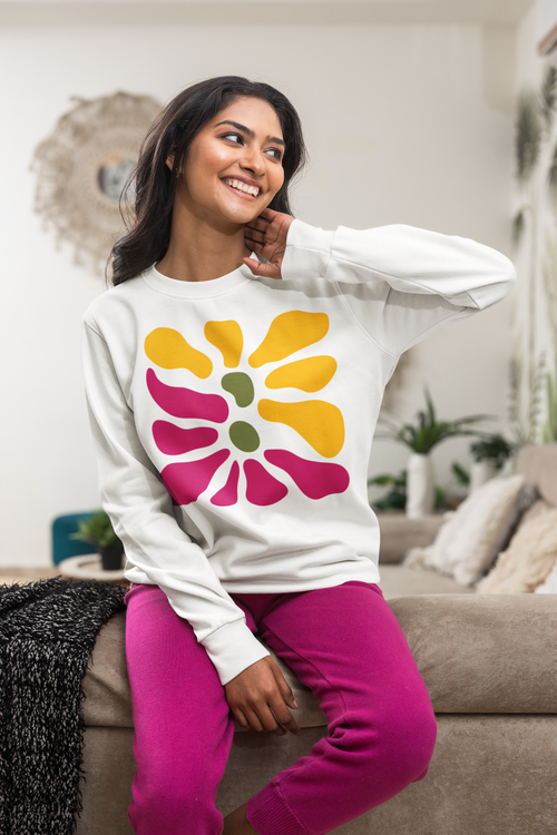 A woman sitting on a beige couch wearing a white crewneck sweatshirt with a retro multi-colored flower graphic and bright pink sweatpants in a cozy living room setting.