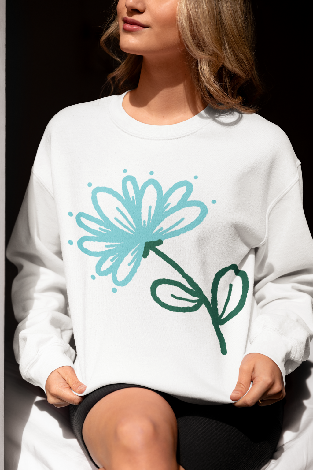 A woman wearing a white crewneck sweatshirt with a stylized blue and green flower graphic on the front, set against a dark background.