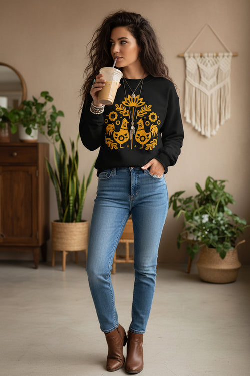 A woman wearing a black crewneck sweatshirt with a yellow folk-art graphic of two roosters and flowers, paired with gray denim shorts and holding an iced coffee.