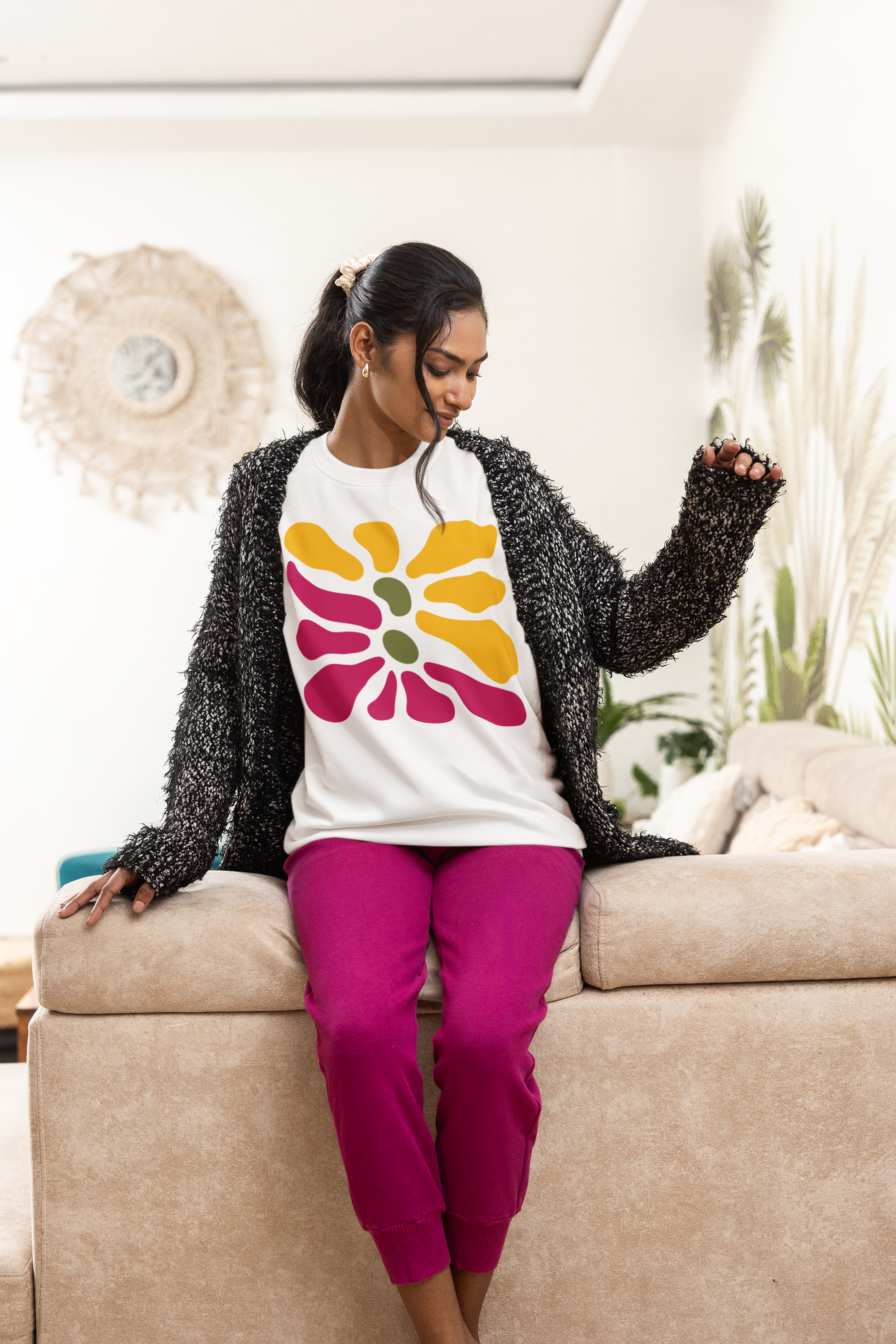 A woman sitting on a beige couch wearing a white crewneck sweatshirt with a colorful flower graphic, a black fuzzy cardigan, and bright pink sweatpants in a living room setting.