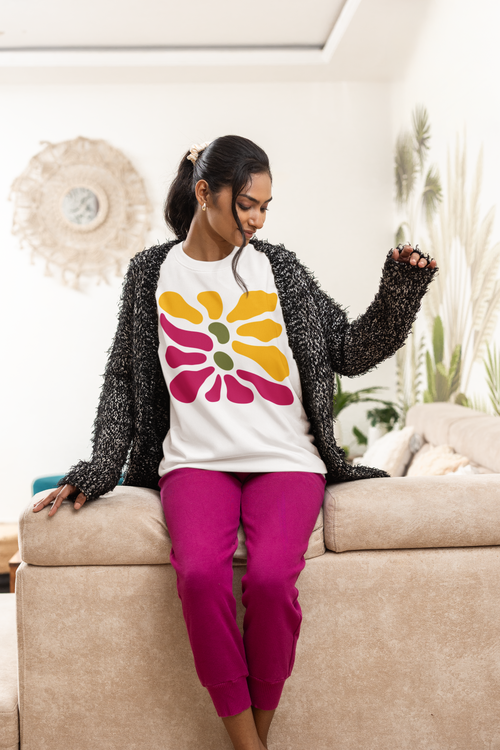 A woman sitting on a beige couch wearing a white crewneck sweatshirt with a colorful flower graphic, a black fuzzy cardigan, and bright pink sweatpants in a living room setting.