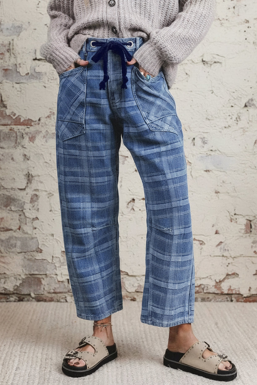 A person wearing blue plaid cropped pants with a dark blue drawstring waist, a gray knit sweater, and beige slide sandals, standing against a plain light wall.