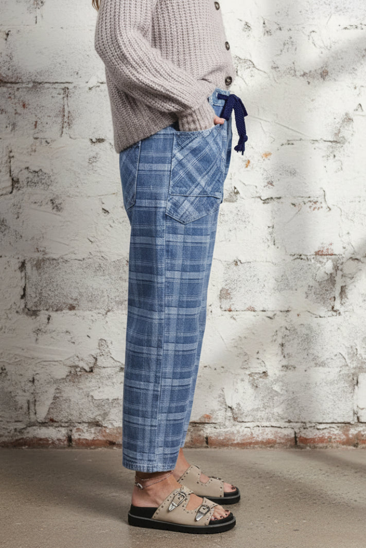 A person wearing blue plaid cropped pants with a dark blue drawstring, a light gray knit cardigan, and beige slide sandals, standing against a plain off-white wall.