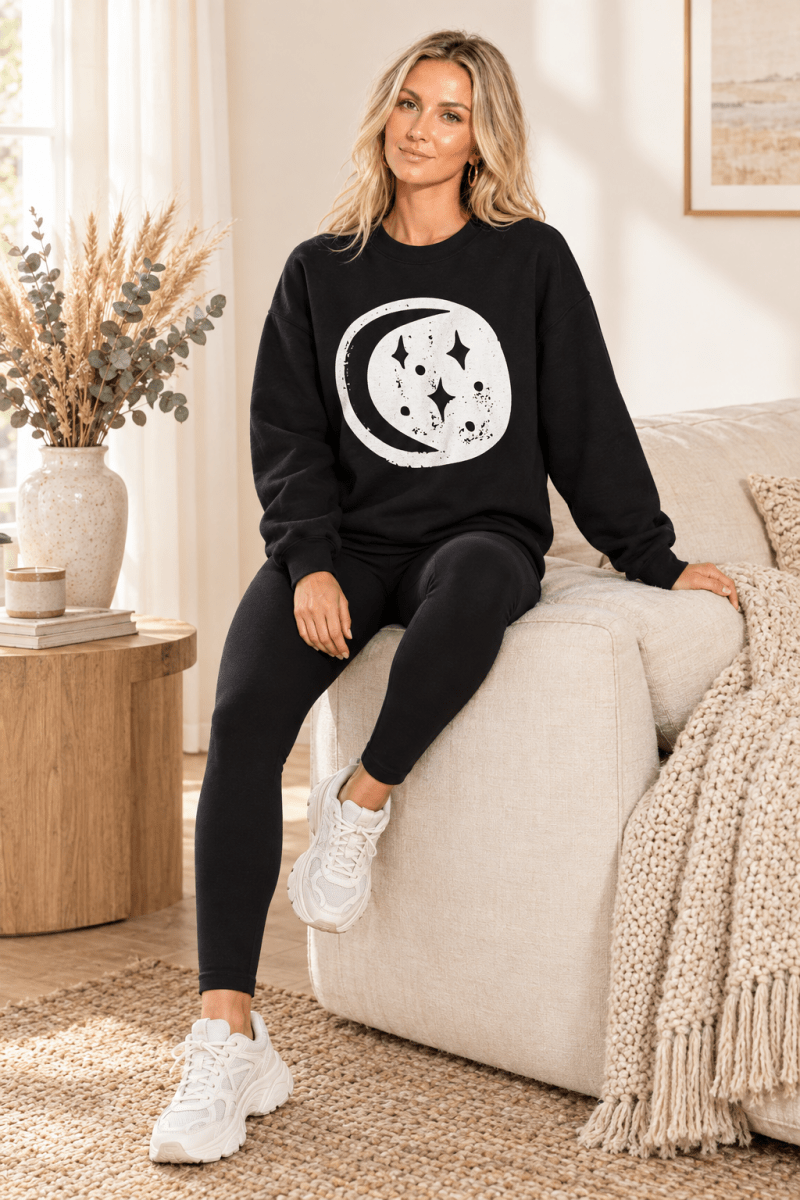 The Doodled Flower Boutique black moon and stars crewneck sweatshirt styled with black leggings and chunky white sneakers in a cream living room.