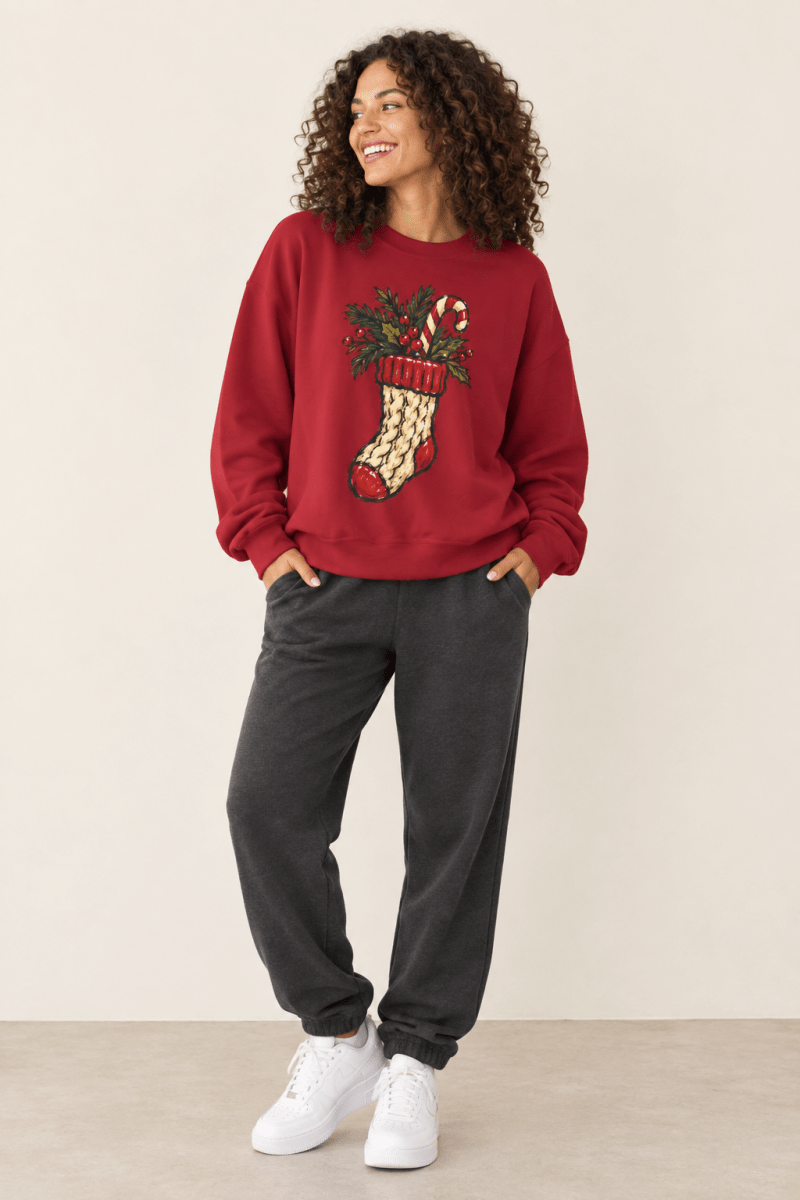 The Doodled Flower Boutique cardinal red crewneck sweatshirt featuring a cable knit Christmas stocking with greenery and a candy cane graphic, worn by a model.