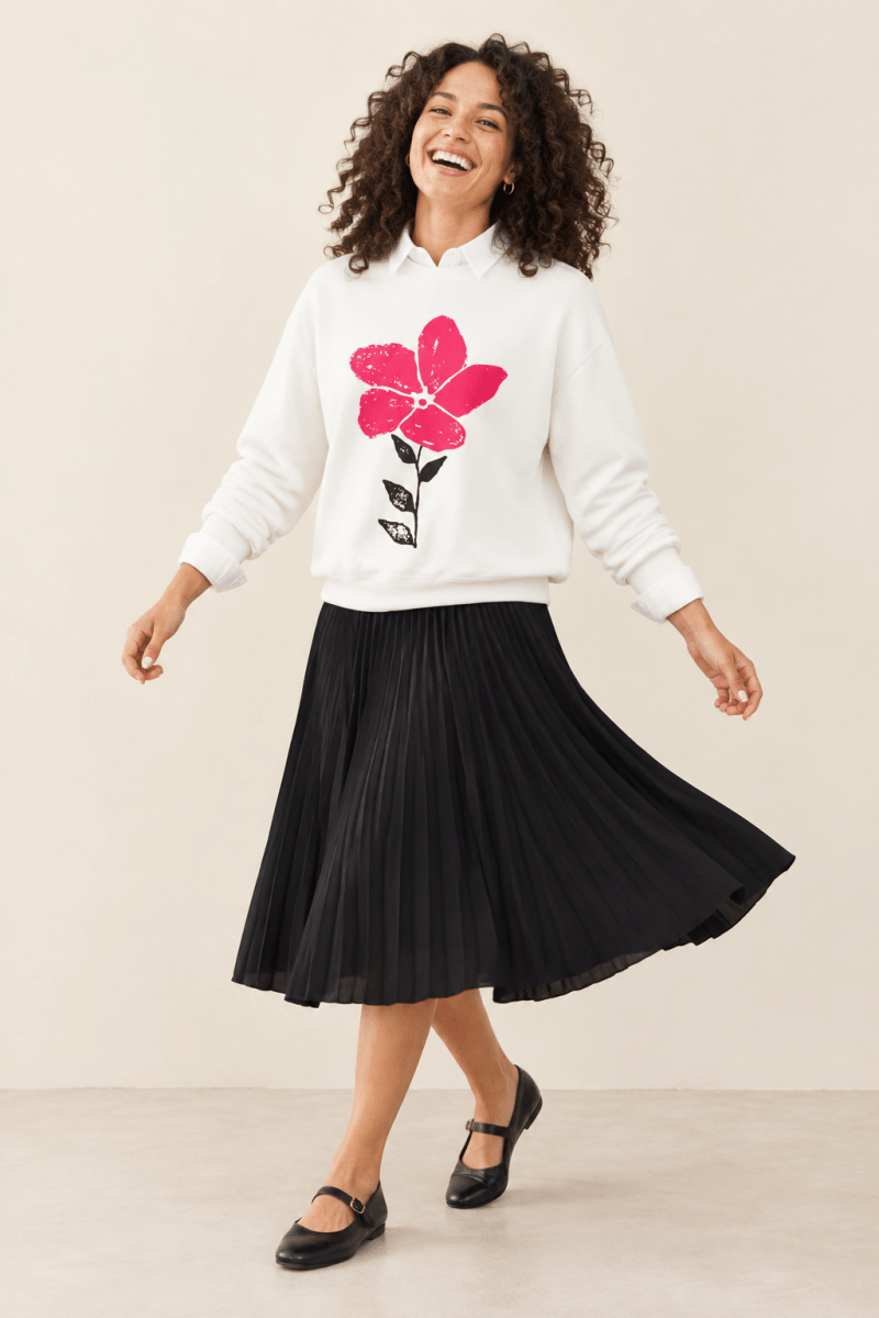 The Doodled Flower Boutique white pink bloom doodle crewneck sweatshirt layered over a white collared shirt with a black pleated midi skirt and mary jane flats.