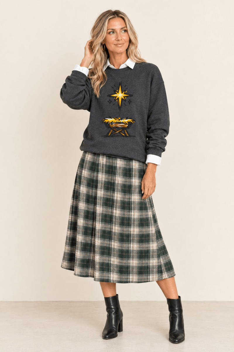 The Doodled Flower Boutique dark heather star of Bethlehem crewneck sweatshirt layered over a white collared shirt with a green plaid midi skirt and black ankle boots.