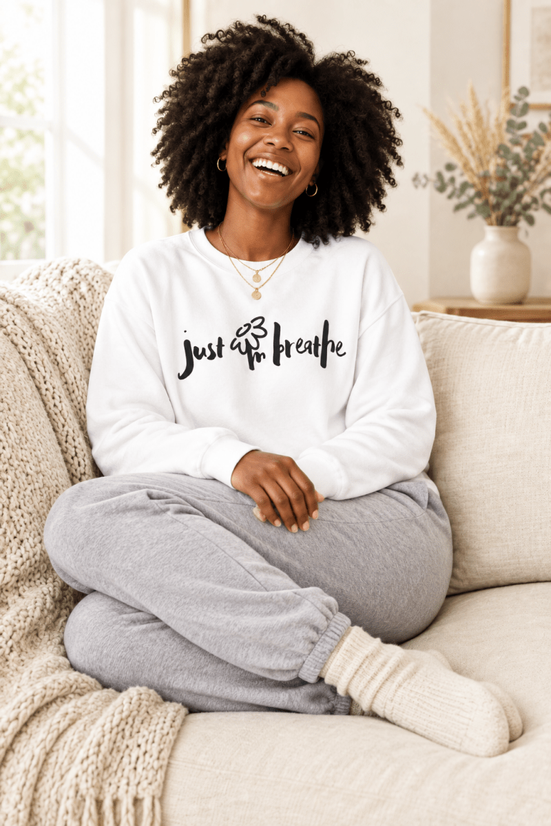 The Doodled Flower Boutique white just breathe crewneck sweatshirt styled with grey joggers on a cozy cream sofa.