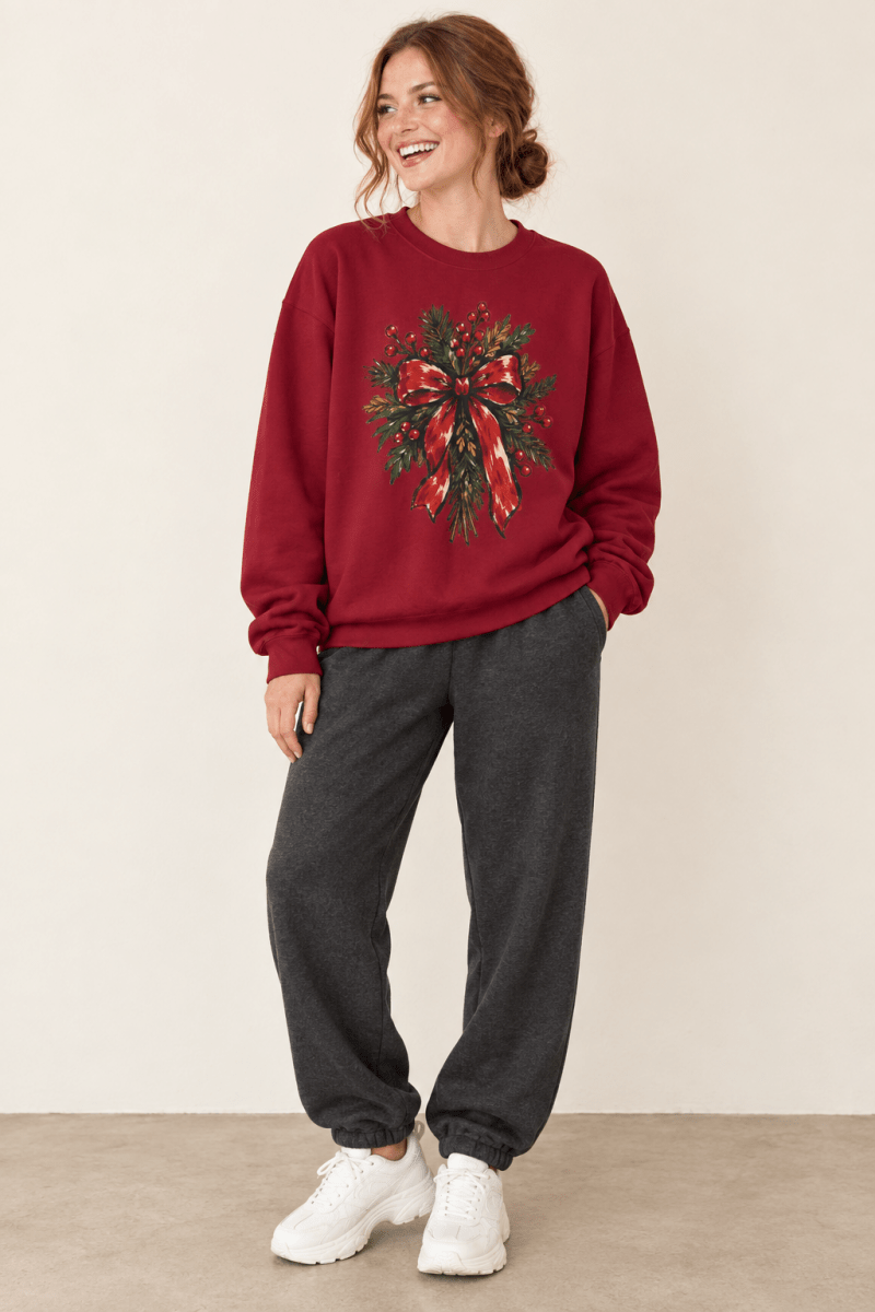 The Doodled Flower Boutique cardinal red crewneck sweatshirt featuring a Christmas wreath with red berries and a red bow graphic, worn by a model.