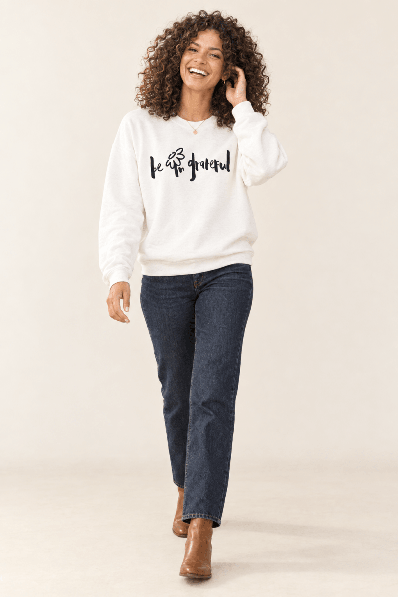 The Doodled Flower Boutique white be grateful crewneck sweatshirt worn by a model with dark wash jeans and cognac ankle boots.