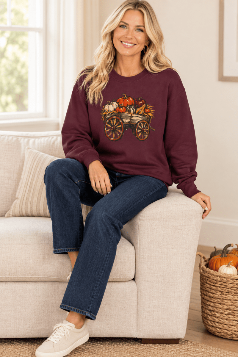 The Doodled Flower Boutique maroon crewneck sweatshirt featuring a rustic pumpkin wagon graphic, worn by a model in a cozy living room