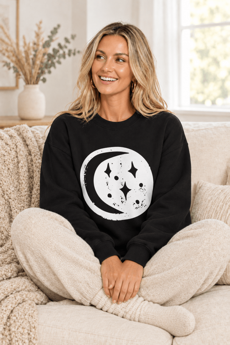The Doodled Flower Boutique black moon and stars crewneck sweatshirt styled with oatmeal knit joggers on a cozy cream sofa.