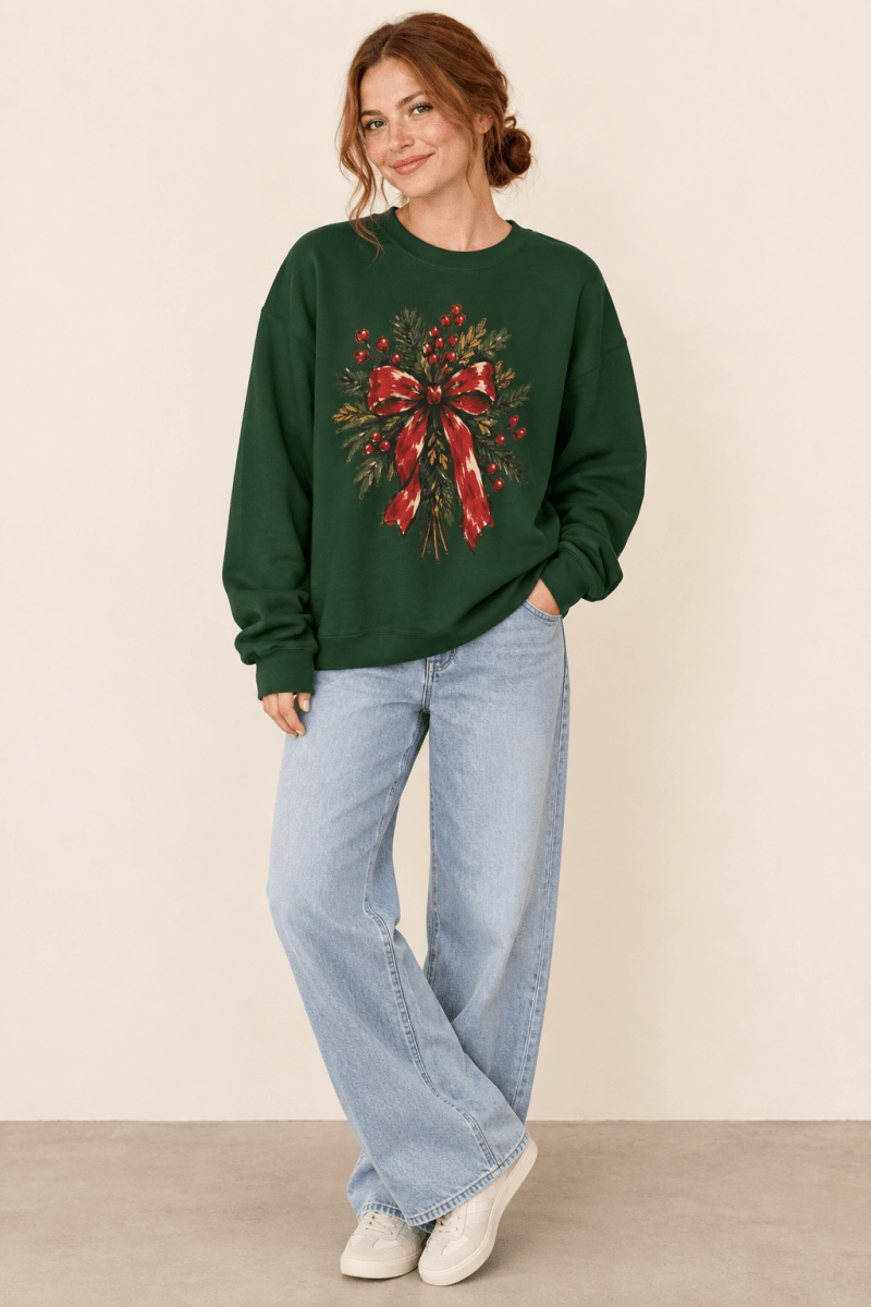 The Doodled Flower Boutique forest green crewneck sweatshirt featuring a Christmas wreath with red berries and a red bow graphic, worn by a model.