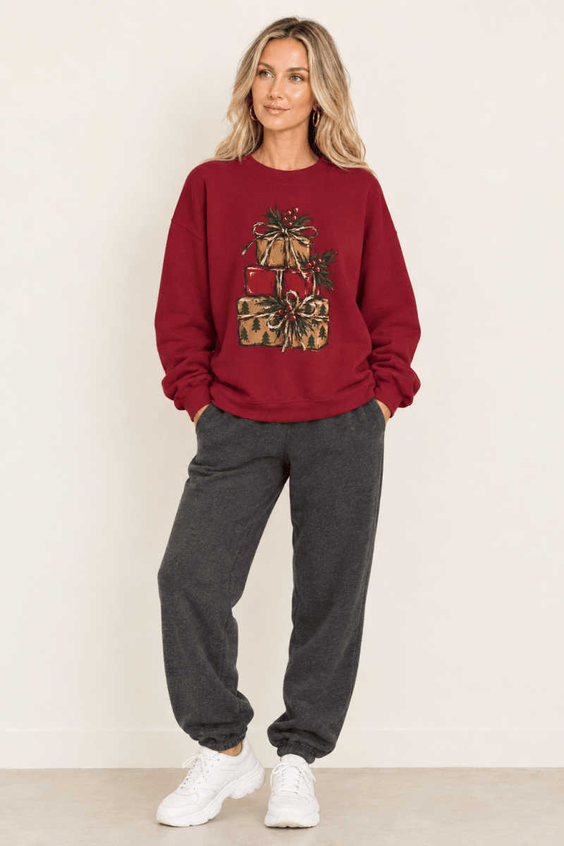 The Doodled Flower Boutique cardinal red crewneck sweatshirt featuring a stacked presents graphic with evergreen sprigs and red berries, worn by a model.