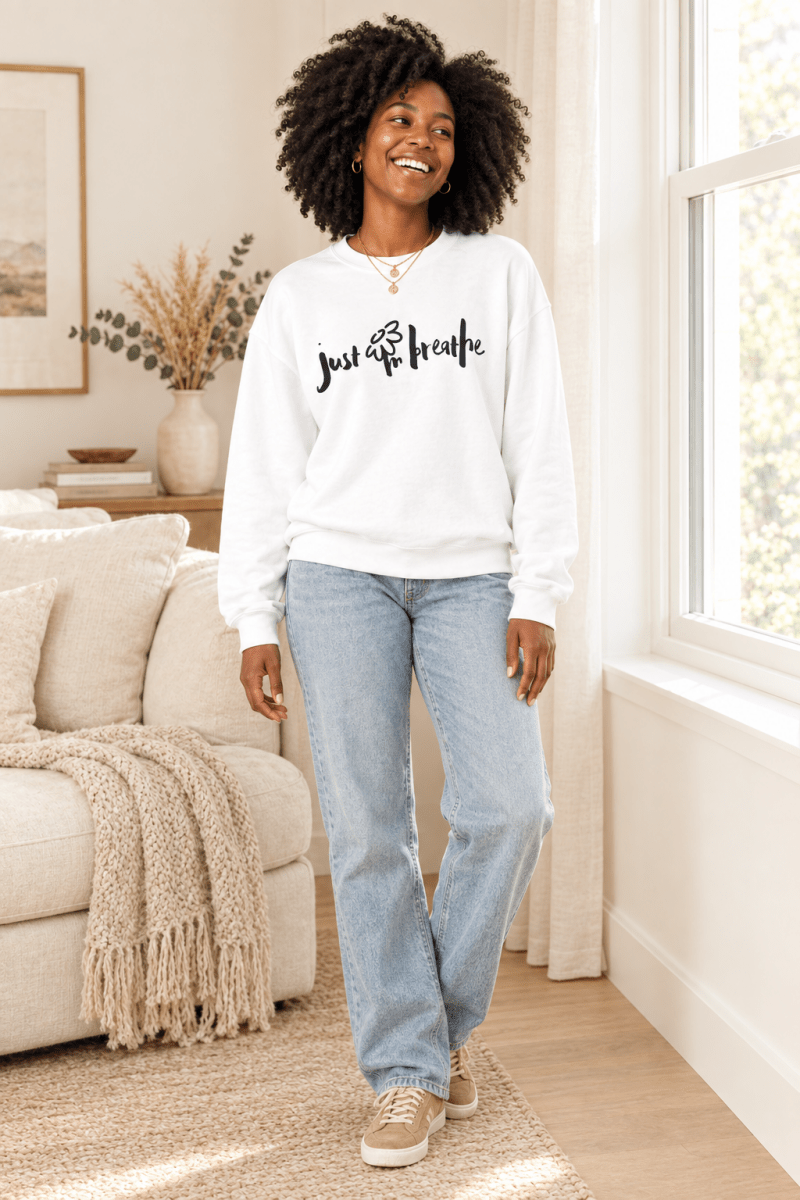 The Doodled Flower Boutique white just breathe crewneck sweatshirt styled with light wash jeans and suede sneakers in a cream living room.