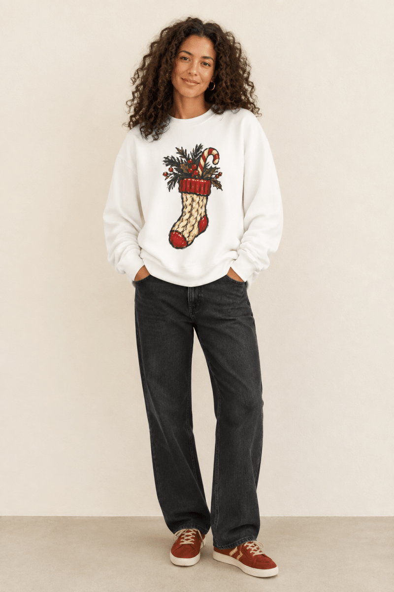 The Doodled Flower Boutique white crewneck sweatshirt featuring a cable knit Christmas stocking with greenery and a candy cane graphic, worn by a model.