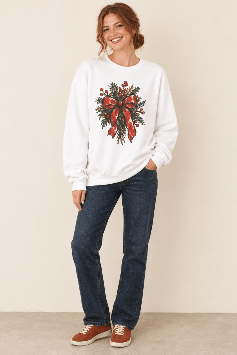 The Doodled Flower Boutique white crewneck sweatshirt featuring a Christmas wreath with red berries and a red bow graphic, worn by a model.
