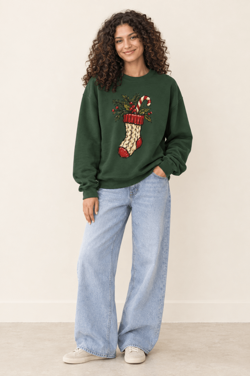 The Doodled Flower Boutique forest green crewneck sweatshirt featuring a cable knit Christmas stocking with greenery and a candy cane graphic, worn by a model.