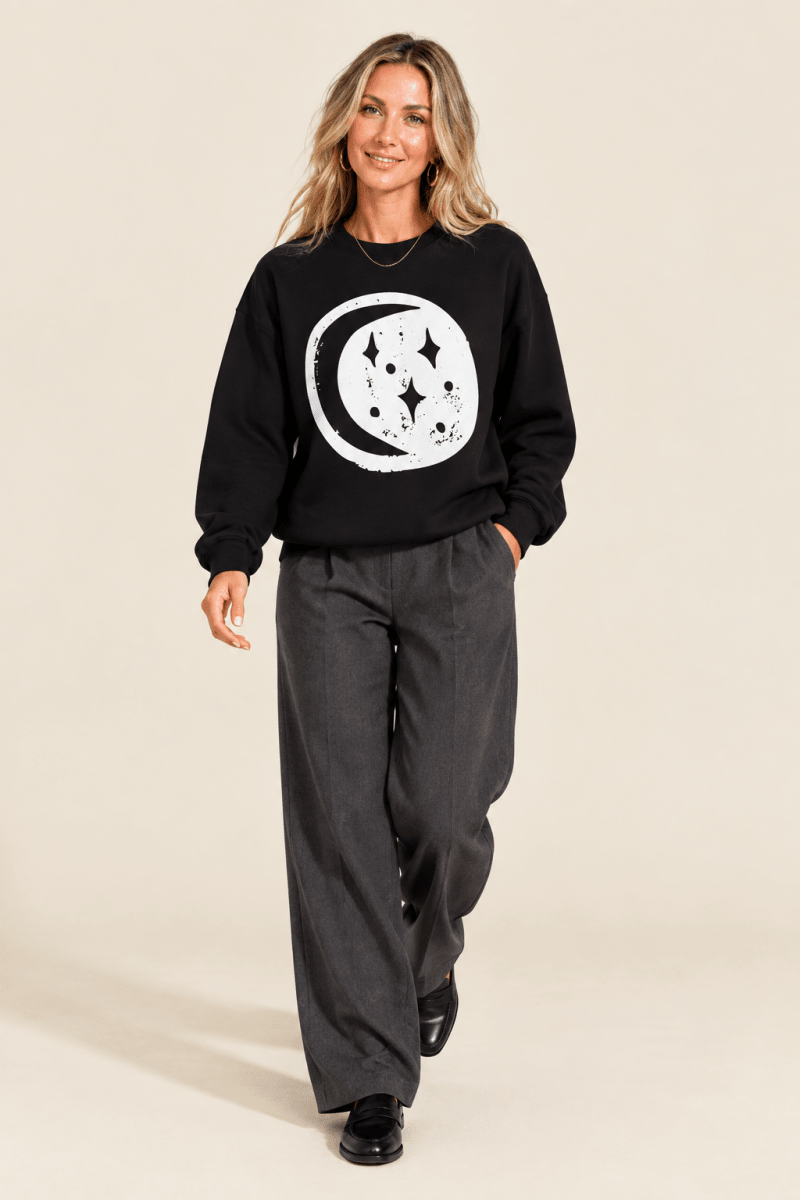 The Doodled Flower Boutique black moon and stars crewneck sweatshirt worn by a model with charcoal wide leg trousers and black loafers.