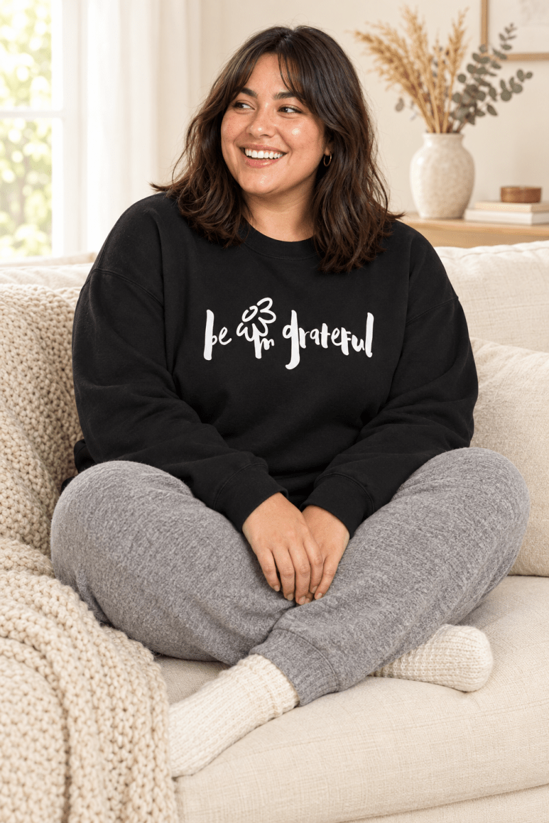 The Doodled Flower Boutique black be grateful crewneck sweatshirt styled with grey knit joggers on a cozy cream sofa.