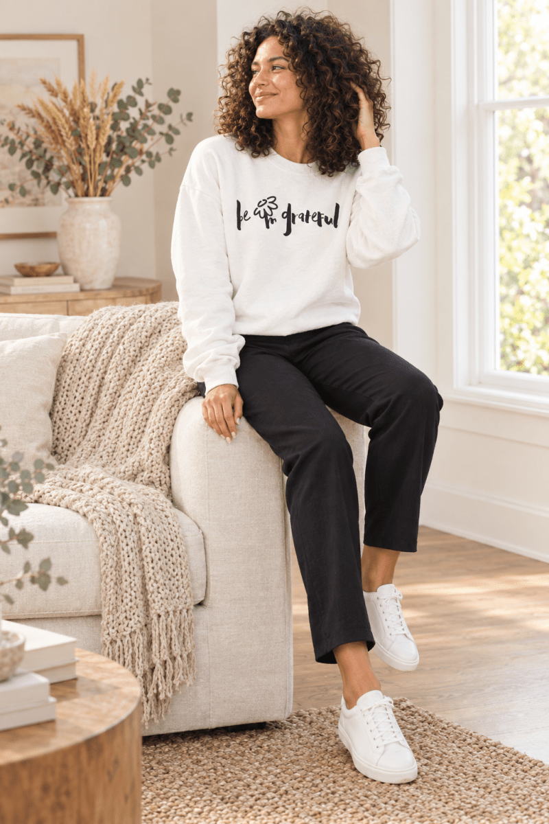 The Doodled Flower Boutique white be grateful crewneck sweatshirt styled with black ankle trousers and white sneakers in a cream living room.