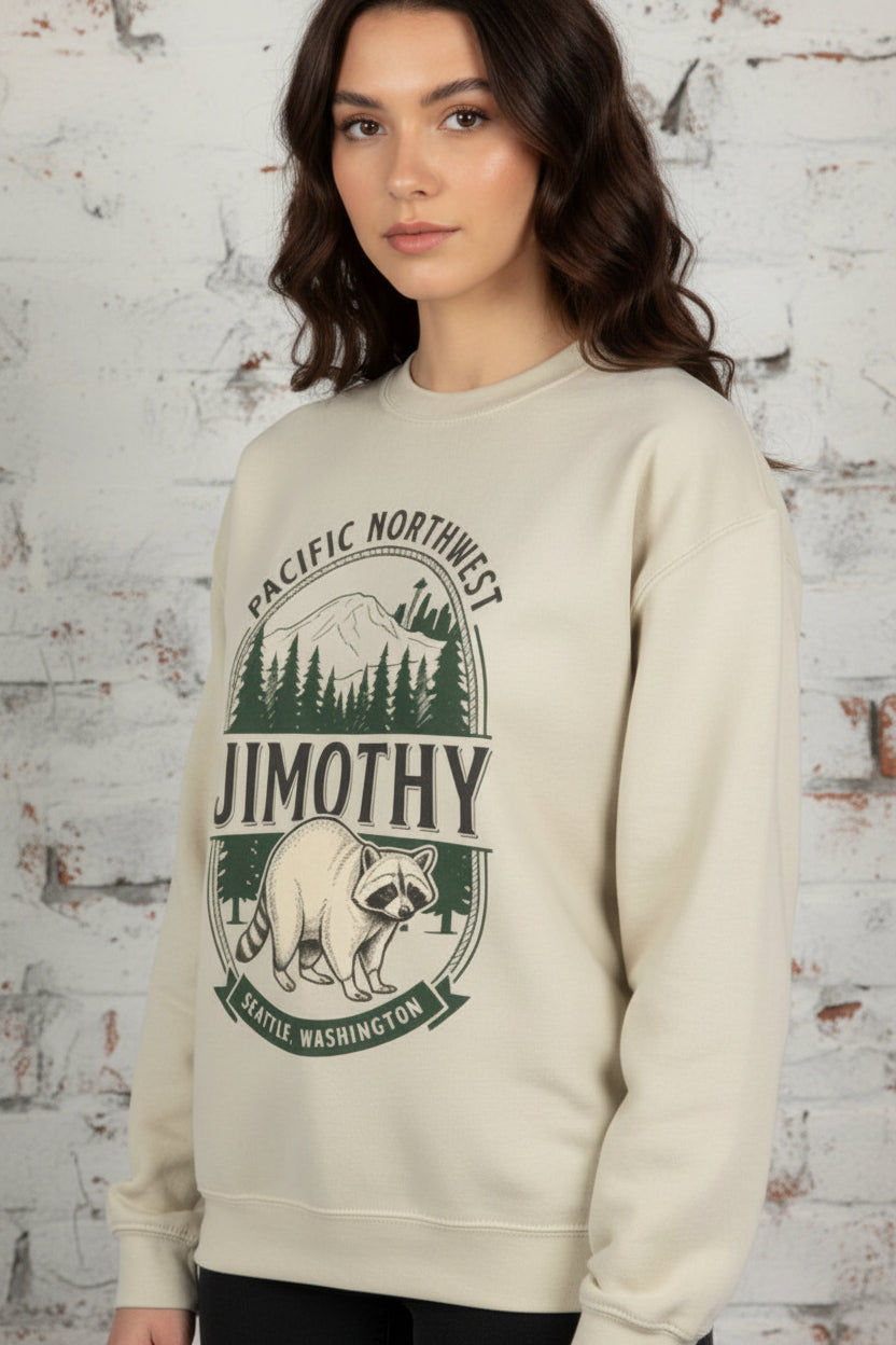 A woman wearing a beige crewneck sweatshirt with a green and black 'Pacific Northwest Jimmythy Seattle, Washington' raccoon graphic, paired with gray denim shorts and holding an iced coffee.
