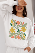 A woman wearing a white sweatshirt with a colorful retro floral pattern on the front, paired with white sweatpants. She is smiling and covering one eye with her hand in a bright indoor setting.