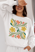 A woman wearing a white sweatshirt with a colorful retro floral pattern on the front, paired with white sweatpants. She is smiling and covering one eye with her hand in a bright indoor setting.