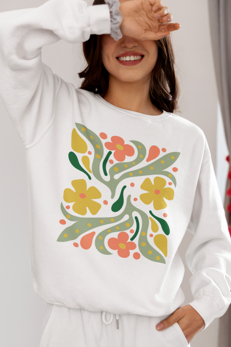 A woman wearing a white sweatshirt with a colorful retro floral pattern on the front, paired with white sweatpants. She is smiling and covering one eye with her hand in a bright indoor setting.