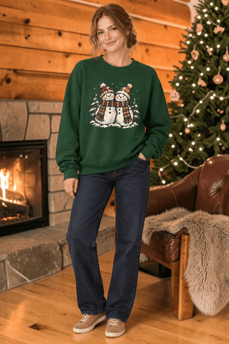 The Doodled Flower Boutique Forest Green crewneck sweatshirt featuring two snowpeople in plaid scarves with pine trees and falling snow, worn by a model.