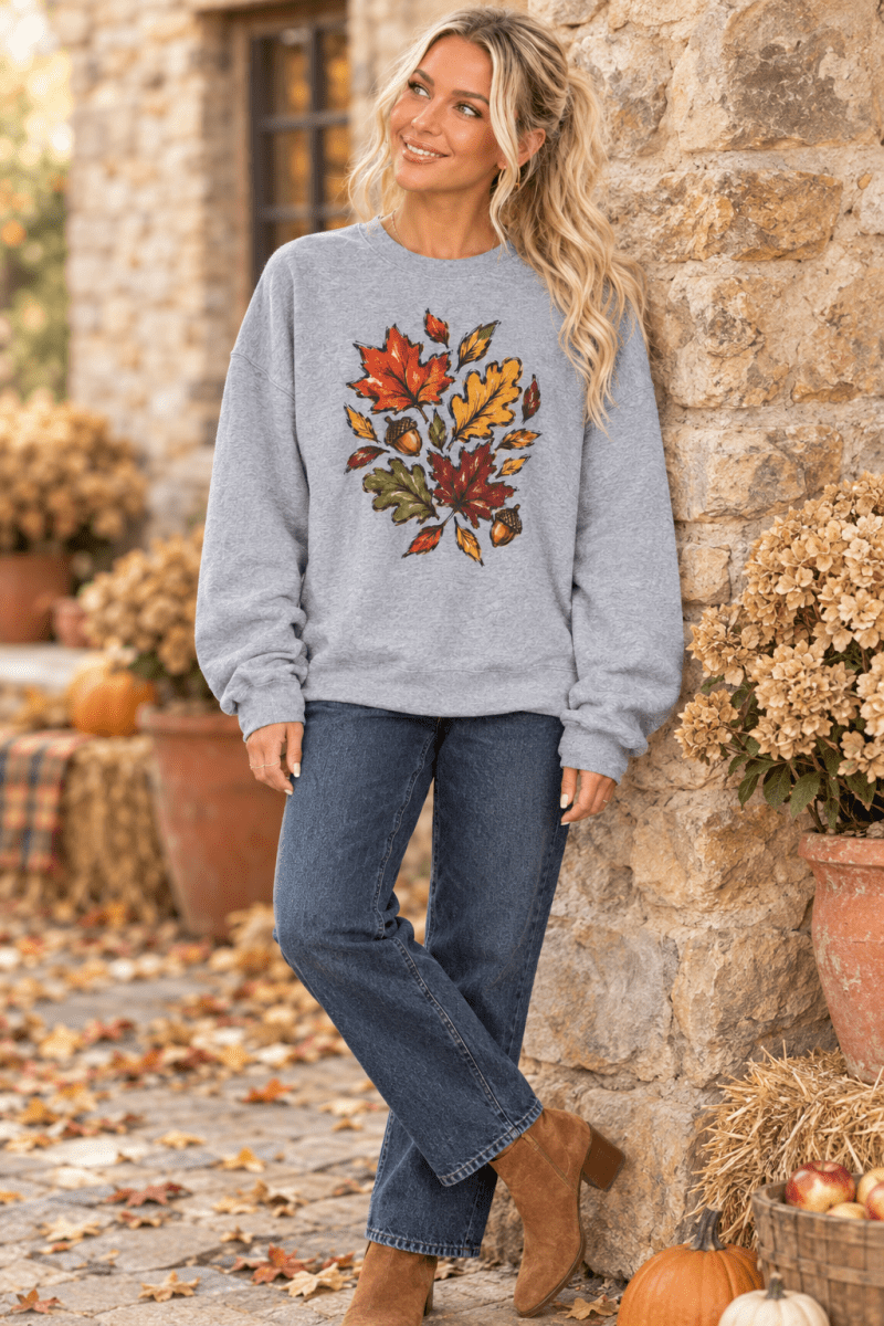 The Doodled Flower Boutique grey crewneck sweatshirt featuring a colorful autumn leaf and acorn print on a woman