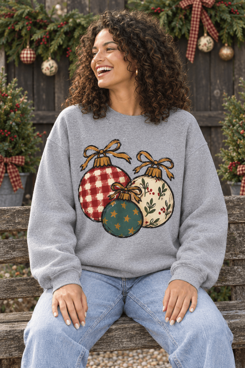 Woman wearing a grey The Doodled Flower Boutique crewneck sweatshirt with hand-drawn Christmas ornament illustrations.