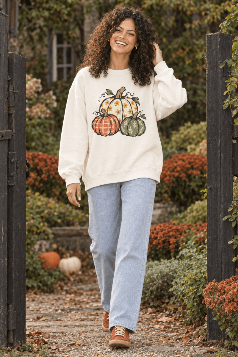 The Doodled Flower Boutique ivory crewneck sweatshirt featuring a hand-drawn pumpkin trio print worn by a smiling woman