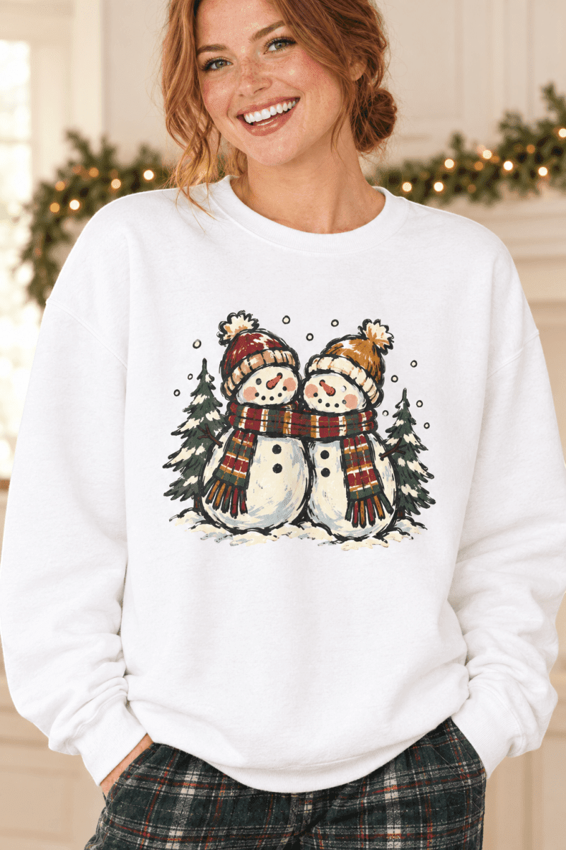 The Doodled Flower Boutique White crewneck sweatshirt featuring two snowpeople in plaid scarves with pine trees and falling snow, worn by a model.
